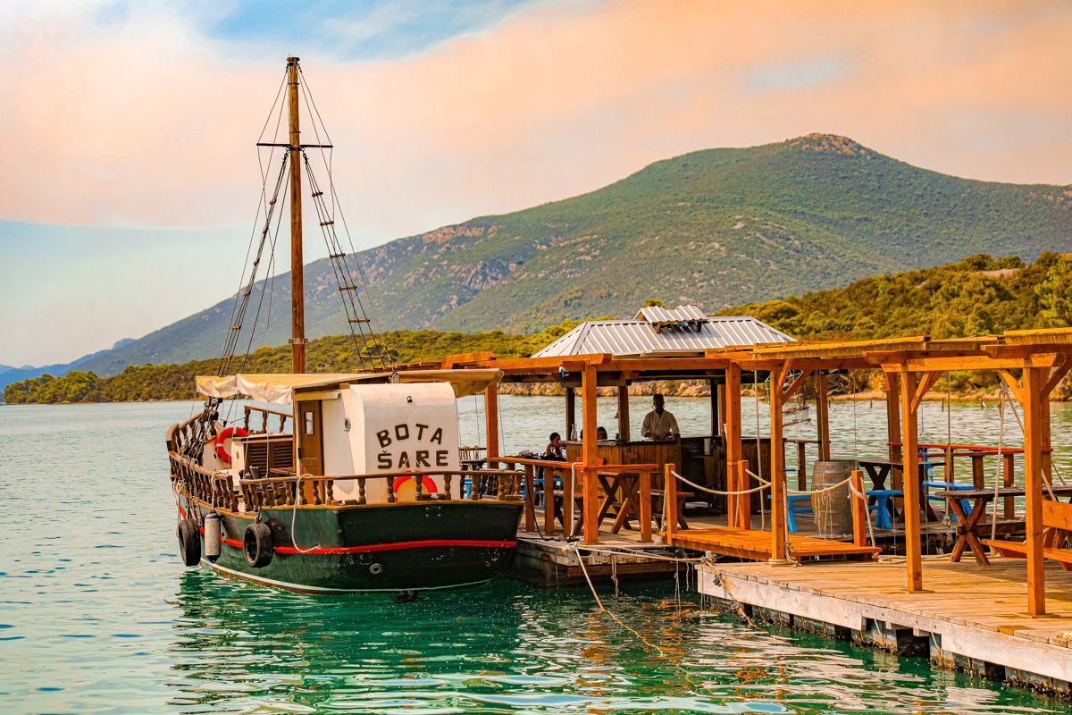 A wooden boat named "Bota Sare" docked at a rustic pier with people under a shaded area, surrounded by clear turquoise water and scenic mountains in the background, awaits your Oyster Day Trip in beautiful Ston, Croatia.