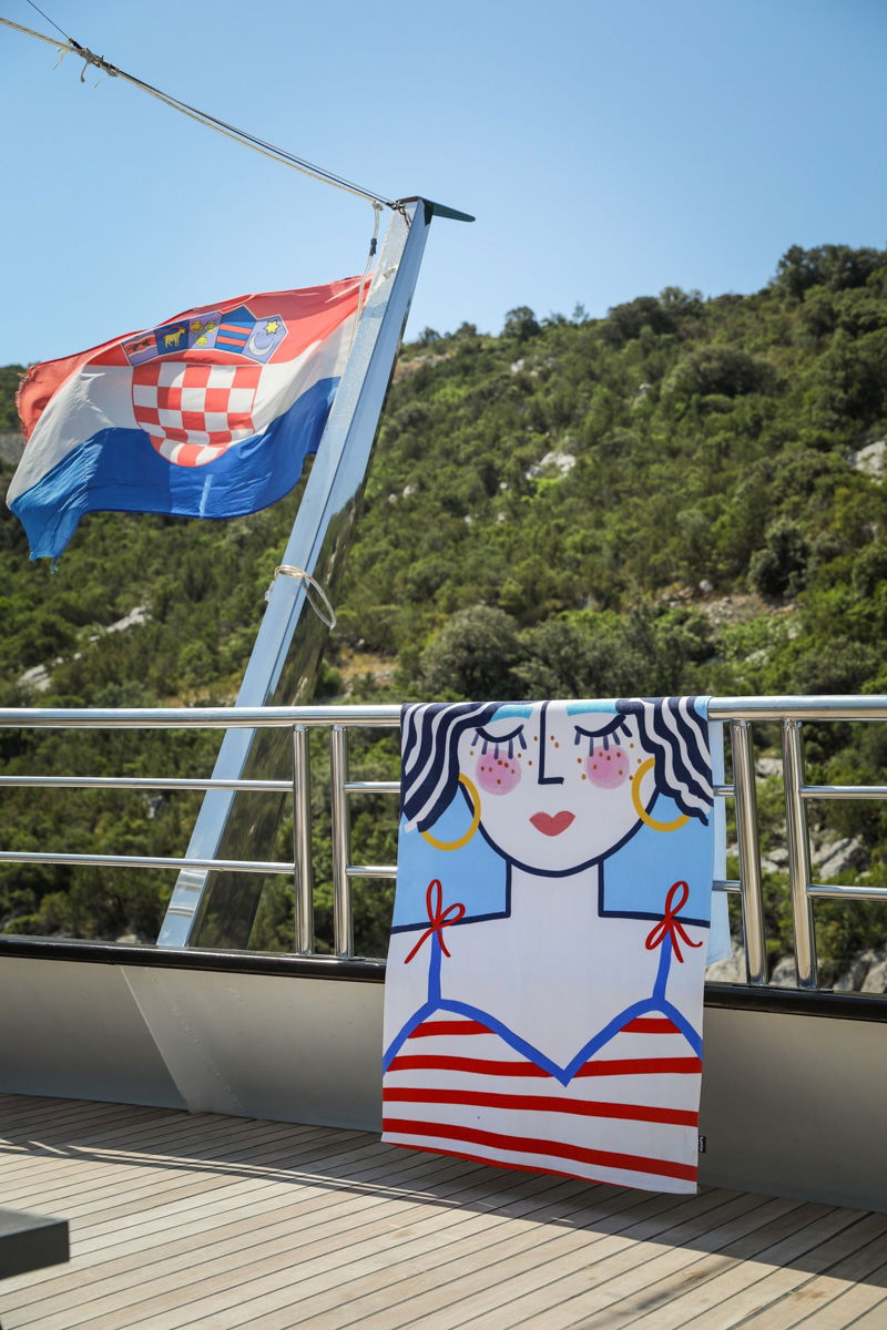 A Croatian flag and a colorful towel with a cartoon illustration of a woman's face hang on the railing of a boat, with a backdrop of trees and hillside—a perfect setup for swimming in the Adriatic Sea.